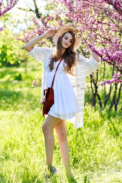 Niesamowita młoda atrakcyjna kobieta w białej lekkiej sukience z długimi włosami, w kapeluszu spaceru w słonecznym ogrodzie w okresie letnim. Kwitnąca sakura, jasne kolory, patrząc w kamerę, stylowy wrażliwy model, zrelaksuj się