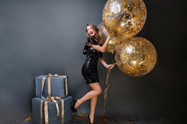 Niesamowita modna młoda kobieta na obcasach, w czarnej luksusowej sukience z dużymi balonami wypełnionymi złotymi błyskotkami. Prezenty, urodziny, świętowanie, uśmiech, wyrażanie pozytywności.