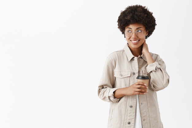 Nieśmiała afrykańska amerykańska pracownica biurowa w koszuli i okularach dotykająca szyi, patrząc w lewo z uśmiechem i trzymając papierowy kubek