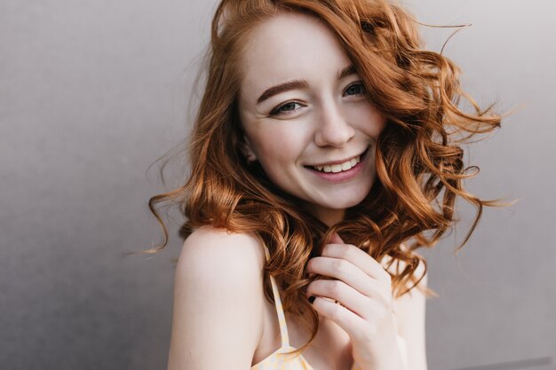 Nieśmiała, czarująca dziewczyna z czerwonymi włosami uśmiechnięta na szarej ścianie. Zdjęcie wspaniałej kręconej modelki korzystającej z dobrego dnia.