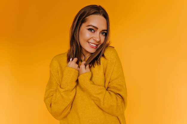Nieśmiała Europejska kobieta pozuje z uśmiechem na pomarańczowej ścianie. Atrakcyjna dziewczyna w żółtym swetrze bawi się.