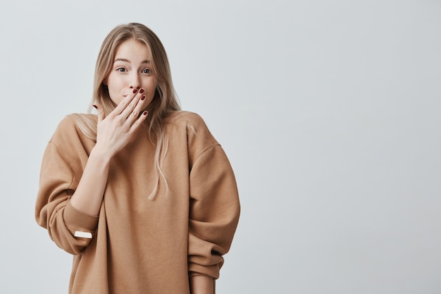 Bezpłatne zdjęcie nieśmiała młoda kobieta. close-up portret kobiety z blond włosami wyglądający na zawstydzonego lub nieśmiałego, będąc w pełnym niedowierzaniu, zszokowany wiadomościami, chowając twarz za rękę, na białym tle.