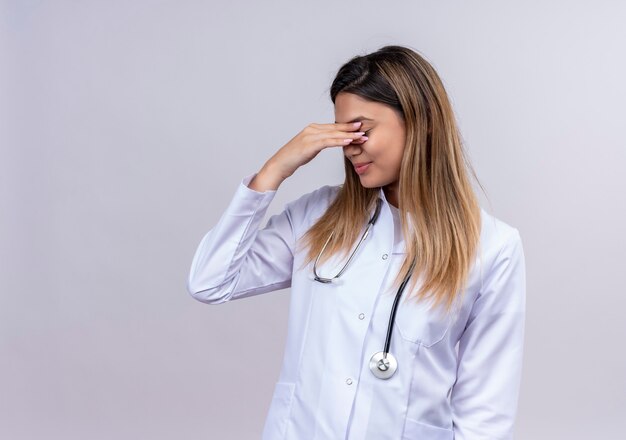 Nieśmiała młoda piękna kobieta lekarz ubrany w biały fartuch ze stetoskopem obejmującym oczy ręką