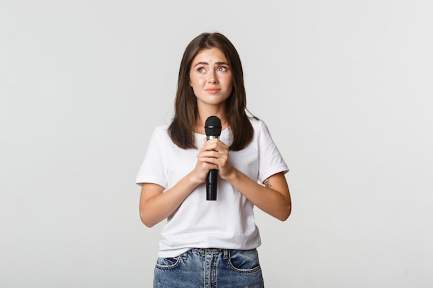 Nieśmiała śliczna brunetka dziewczyna, która boi się śpiewać publicznie, stoi z mikrofonem i wygląda na zdenerwowaną.