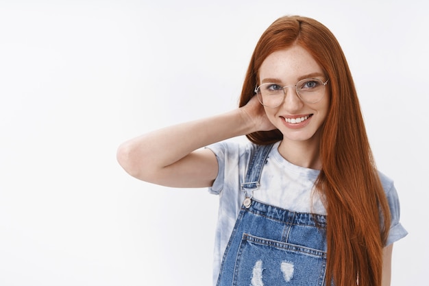 Nieśmiała śliczna rudowłosa studentka długie naturalne rude włosy niebieskie oczy, dotyk szyi skromny uśmiech, rumieniąc się zalotna rozmowa, stoisko biała ściana wesoły entuzjastyczny nastrój