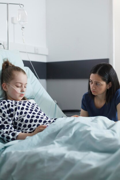 Niespokojna matka opiekuńcza odwiedzająca hospitalizowaną chorą małą dziewczynkę, siedząc w pokoju na oddziale kliniki pediatrycznej. Przemyślany, zmartwiony dorosły wyglądający na zaniepokojonego, wspierając chorą córkę w szpitalnym łóżku.