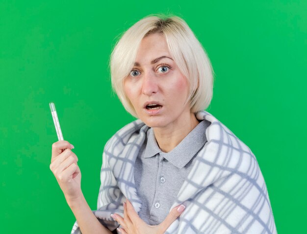Niespokojna młoda blondynka chora kobieta owinięta w kratę trzyma termometr patrząc z przodu na białym tle na zielonej ścianie