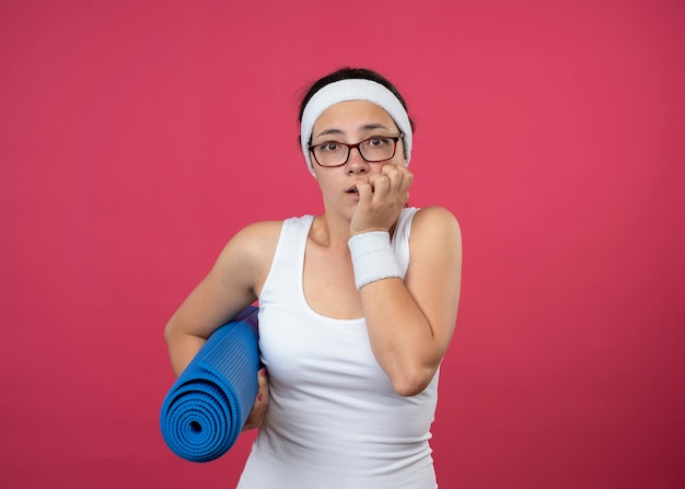 Niespokojna młoda kobieta sportowa w okularach optycznych z opaską na głowę i opaską na nadgarstek gryzie paznokcie i trzyma matę sportową na różowej ścianie