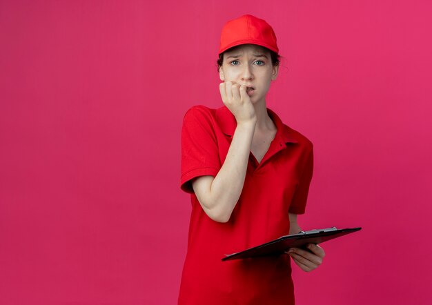 Niespokojna młoda ładna dziewczyna w czerwonym mundurze i czapce trzymająca schowek, patrząc na kamerę i gryzące palce na białym tle na szkarłatnym tle z miejsca na kopię
