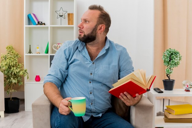 Niespokojny dorosły mężczyzna słowiański siedzi na fotelu, trzymając książkę i kubek, patrząc z boku w salonie