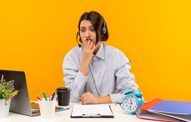 Niespokojny młoda dziewczyna call center sobie zestaw słuchawkowy siedzi przy biurku patrząc z boku ręką na ustach samodzielnie na pomarańczowo
