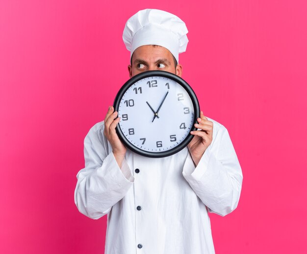 Niespokojny młody kaukaski kucharz w mundurze szefa kuchni i czapce trzymającej zegar patrzący z boku z tyłu na różowej ścianie