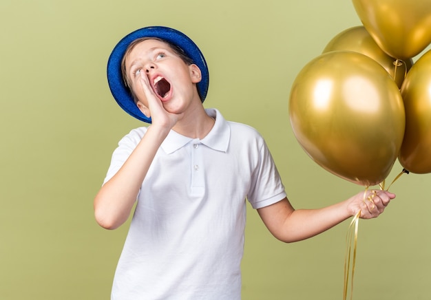 Bezpłatne zdjęcie niespokojny młody słowiański chłopiec w niebieskim kapeluszu imprezowym trzymający balony z helem i trzymający rękę blisko ust wołający kogoś patrzącego w górę odizolowanego na oliwkowozielonej ścianie z kopią przestrzeni