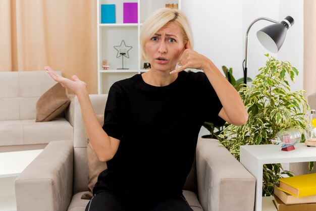 Nieświadoma piękna blondynka Rosjanka siedzi na fotelu, wskazując ręką znak telefonu i trzymając dłoń otwartą w salonie