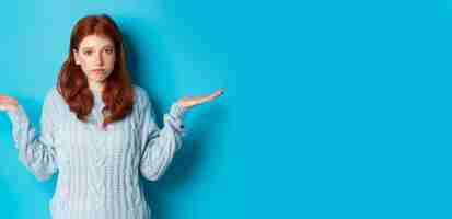 Bezpłatne zdjęcie nieświadoma rudowłosa dziewczyna wzruszająca ramionami i mówiąca „przepraszam”, stojąc zdziwiona na niebieskim tle, nie ma identyfikatora