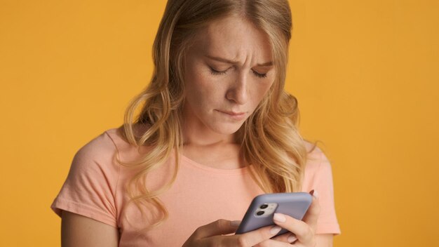 Nieszczęśliwa blond dziewczyna trzymająca smartfona czuje się smutna, ponieważ nie dostała wiadomości od chłopaka wyizolowanego na żółtym tle