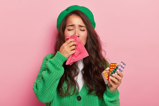 Nieszczęśliwa brunetka cierpi na objawy grypy, wyciera nos chusteczką, jest przeziębiona, trzyma pigułki, nosi zielony sweter i czapkę, odizolowane na różowej ścianie