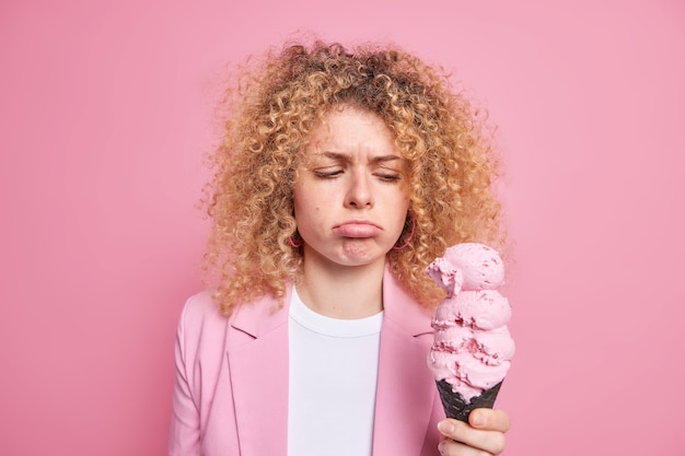 Nieszczęśliwa młoda kobieta z kręconymi włosami torebki usta patrzy smutno na pyszne lody czuje pokusę, by zjeść słodki, smaczny, wysokokaloryczny deser ubrana w formalną kurtkę na białym tle nad różową ścianą