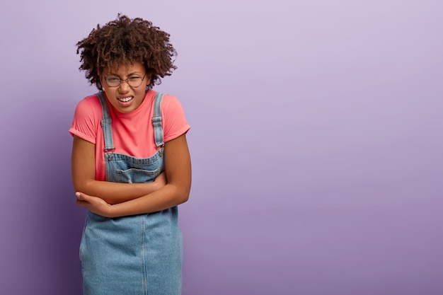 Niezadowolona Afroamerykanka trzyma ręce na brzuchu, zaciska zęby z powodu nieprzyjemnych uczuć, ma dolegliwości lub bóle menstruacyjne, czuje dyskomfort w brzuchu, nosi dżinsową sarafan.