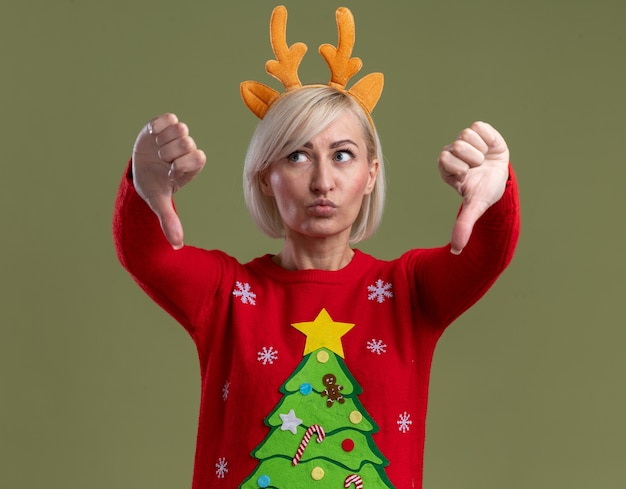 Niezadowolona Blondynka W średnim Wieku Ubrana W świąteczną Opaskę Z Poroża Renifera I świąteczny Sweter Patrząc Z Boku, Pokazując Kciuki W Dół Z Zaciśniętymi Ustami Odizolowanymi Na Oliwkowym Tle