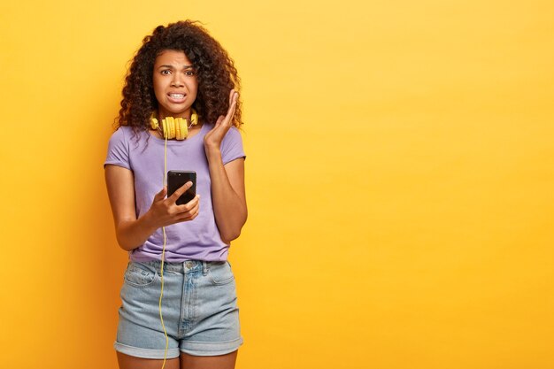 Niezadowolona ciemnoskóra kręcona kobieta słucha obrzydliwej wiadomości głosowej w słuchawkach, trzyma nowoczesny smartfon, nosi fioletową koszulkę i dżinsowe szorty, stoi na żółtej ścianie studia, kopia przestrzeń