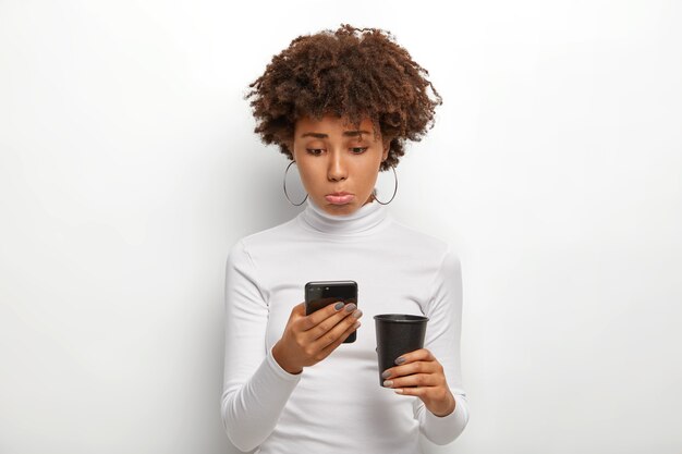 Niezadowolona czarna kobieta z kręconymi włosami, smutno patrzy na telefon