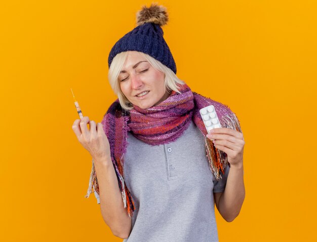 Niezadowolona młoda blondynka chora słowiańska kobieta w czapce zimowej i szaliku trzyma strzykawkę i paczkę pigułek medycznych odizolowanych na pomarańczowej ścianie z miejscem na kopię