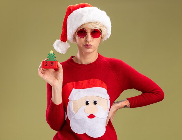 niezadowolona młoda blondynka w świątecznym kapeluszu i mikołajowym świątecznym swetrze w okularach trzymająca rękę w talii trzymająca choinkę zabawka z datą patrzącą na oliwkowo-zielonej ścianie