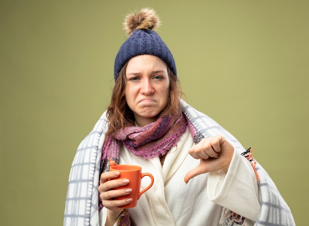 Bezpłatne zdjęcie niezadowolona młoda chora dziewczyna w białej szacie i czapce zimowej z szalikiem owiniętym w kratę trzymająca filiżankę herbaty pokazująca kciuk w dół odizolowany na oliwkowej zieleni