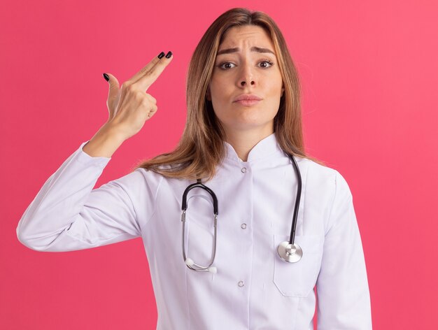 Niezadowolona młoda kobieta lekarz ubrana w szlafrok medyczny ze stetoskopem pokazujący gest pistoletu na białym tle na różowej ścianie