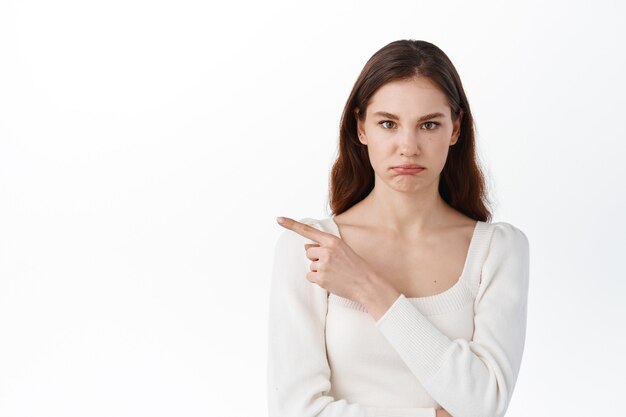 Niezadowolona młoda kobieta wskazująca palcem wskazującym na bok, pokazująca złą promocję, dąsająca się zdenerwowana, wyglądająca na rozczarowaną, stojąca przy białej ścianie