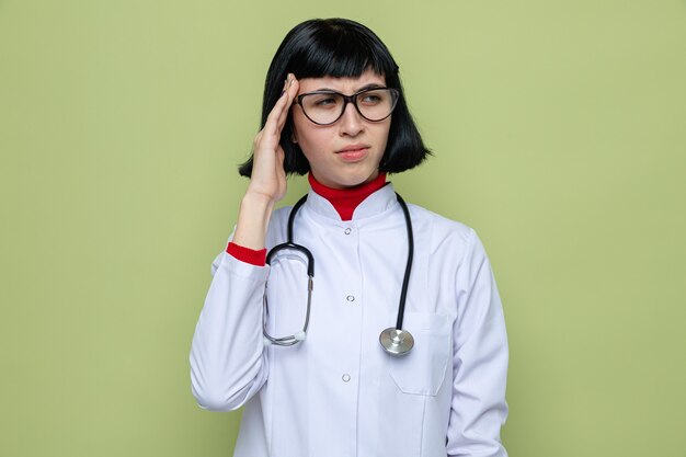 Niezadowolona młoda ładna kaukaska dziewczyna w okularach optycznych w mundurze lekarza ze stetoskopem kładzie rękę na skroni