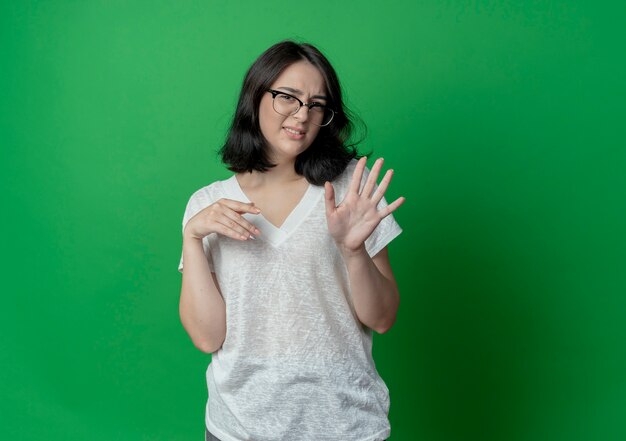 Niezadowolona młoda ładna kaukaska dziewczyna w okularach pokazująca pięć z ręką i trzymając inną w powietrzu na białym tle na zielonym tle z miejsca na kopię