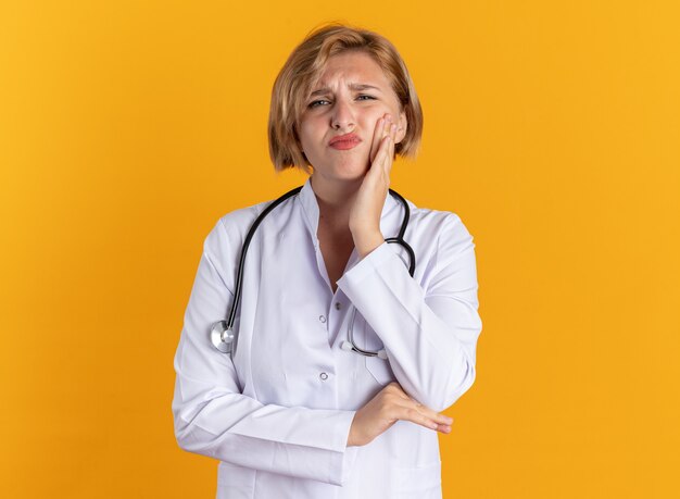 Niezadowolona młoda lekarka nosząca szatę medyczną ze stetoskopem, kładąc rękę na bolącym zębie na białym tle na pomarańczowym tle