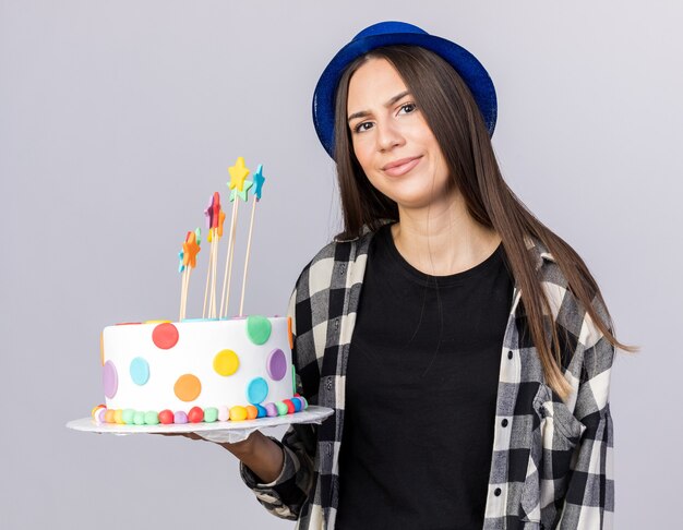 Niezadowolona młoda piękna dziewczyna w kapeluszu imprezowym trzymająca ciasto na białym tle na białej ścianie