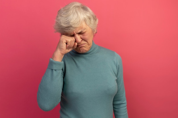 Niezadowolona stara kobieta w niebieskim swetrze z golfem i okularach przeciwsłonecznych wycierająca oko z zamkniętymi oczami odizolowanymi na różowej ścianie z kopią przestrzeni