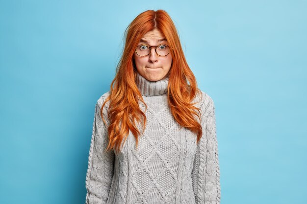 Niezadowolona zdziwiona ruda młoda kobieta zaciska usta i wygląda na zawstydzoną, ma zszokowany wyraz twarzy ubrany w sweter i okulary.