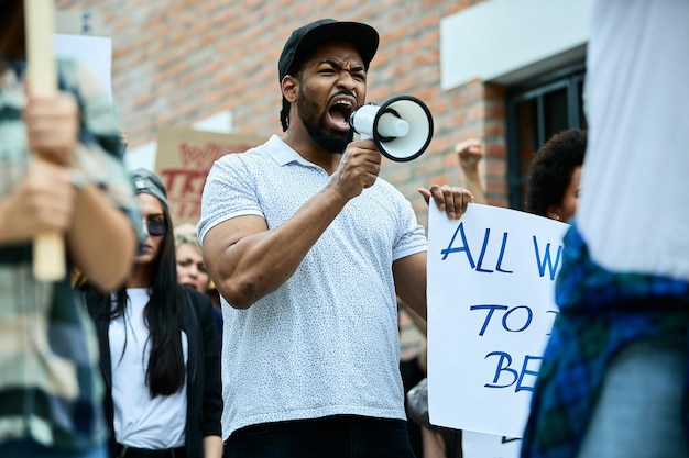 Bezpłatne zdjęcie niezadowolony afroamerykanin krzyczy przez megafon podczas demonstracji w obronie praw człowieka wśród tłumu ludzi na ulicach
