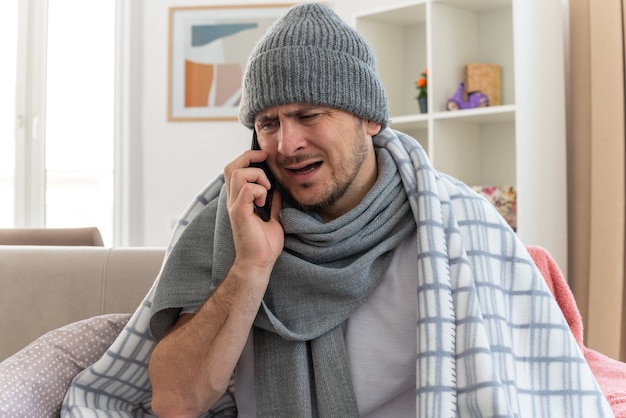 Niezadowolony chory mężczyzna z szalikiem na szyi w czapce zimowej owiniętej w kratę rozmawiający przez telefon siedzący na kanapie w salonie