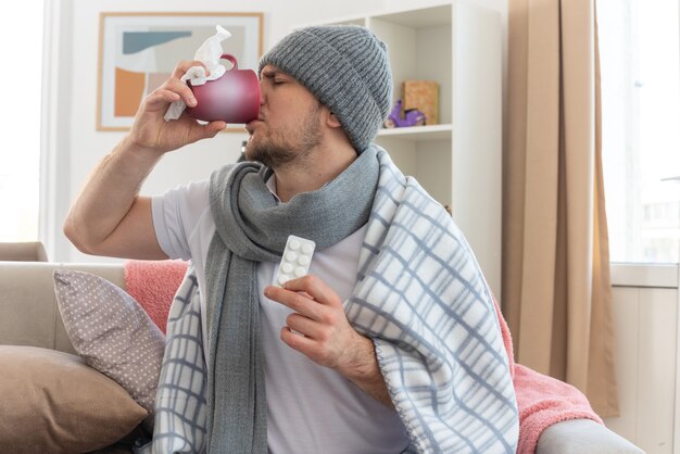 Niezadowolony chory słowiański mężczyzna z szalikiem na szyi w czapce zimowej owiniętej w kratę, trzymający blistry z lekarstwami i pijący z kubka siedzącego na kanapie w salonie