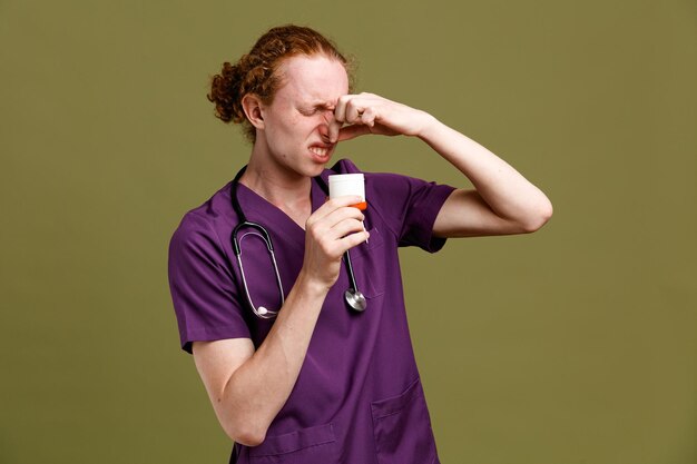 Niezadowolony chwyt nosowy może młody lekarz mężczyzna ubrany w mundur ze stetoskopem na zielonym tle