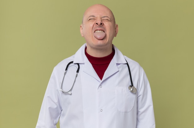 Niezadowolony dorosły słowiański mężczyzna w mundurze lekarza ze stetoskopem wystawia język, stojąc z zamkniętymi oczami