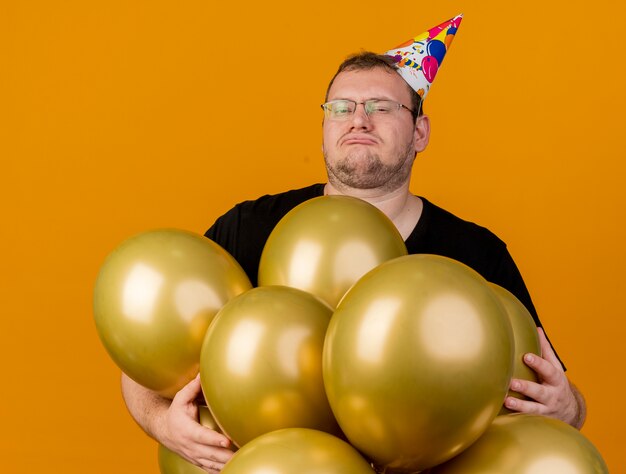 Niezadowolony dorosły słowiański mężczyzna w okularach optycznych noszący czapkę urodzinową z balonami z helem