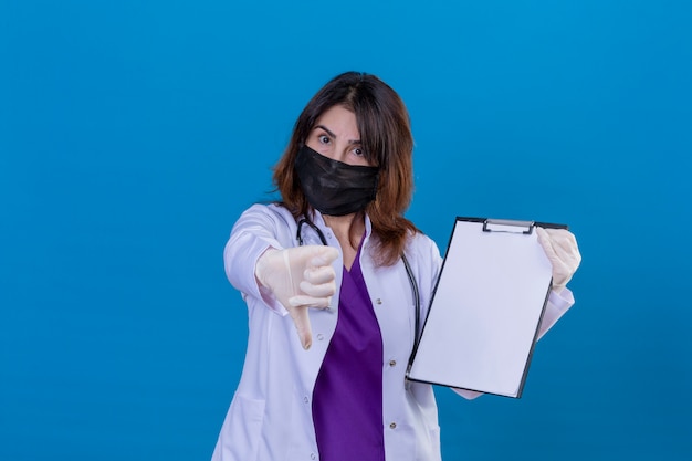 Niezadowolony lekarz w średnim wieku w białym fartuchu w czarnej ochronnej masce na twarz i ze stetoskopem trzymającym notatnik pokazujący kciuk w dół nad niebieską ścianą