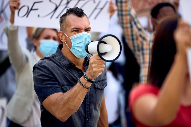 Niezadowolony mężczyzna używający megafonu i krzyczący podczas noszenia maski ochronnej i biorący udział w demonstracji zapobiegającej blokowaniu się na ulicach miasta