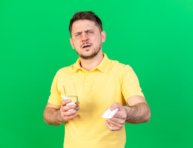 Niezadowolony młody blond chory słowiański mężczyzna trzyma szklankę wody i paczkę tabletek medycznych na białym tle na zielonej ścianie z miejsca na kopię