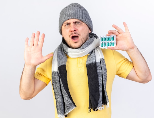 Bezpłatne zdjęcie niezadowolony młody blond chory słowiański mężczyzna w czapce zimowej i szaliku stoi z podniesioną ręką i trzyma paczkę tabletek medycznych na białej ścianie z miejscem na kopię