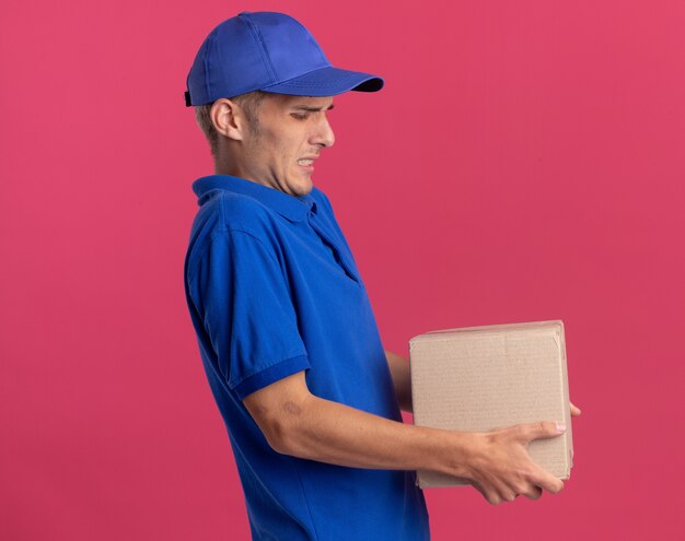 Niezadowolony młody blondyn-dostawca stoi bokiem, trzymając ciężkie kartonowe pudełko