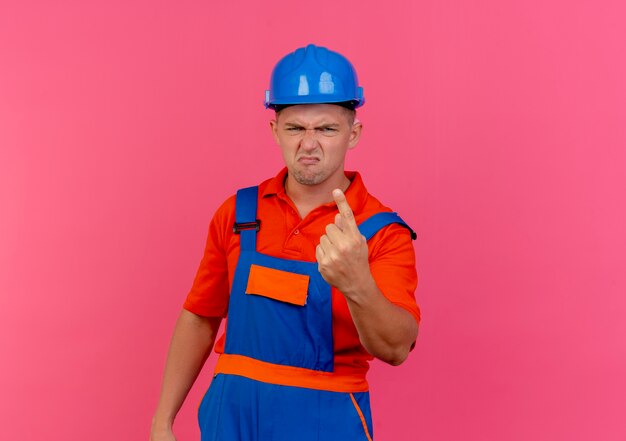 Bezpłatne zdjęcie niezadowolony młody budowniczy mężczyzna w mundurze i hełmie ochronnym, na którym widać jeden na różowo