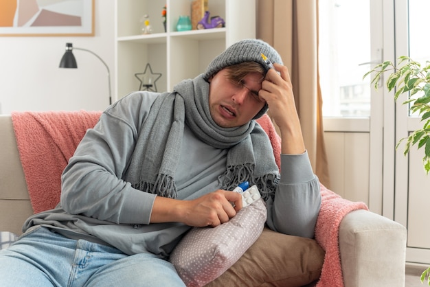 niezadowolony młody chory mężczyzna z szalikiem na szyi w czapce zimowej trzymający blistry strzykawki i lekarstwa oraz termometr siedzący na kanapie w salonie